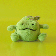 On a green background is a small green stuffed toy frog shaped bag charm.
