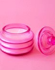 On a pink background is a glass pink jarred candle with a coordinating lid.