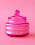 On a pink background is a glass pink jarred candle with a coordinating lid.