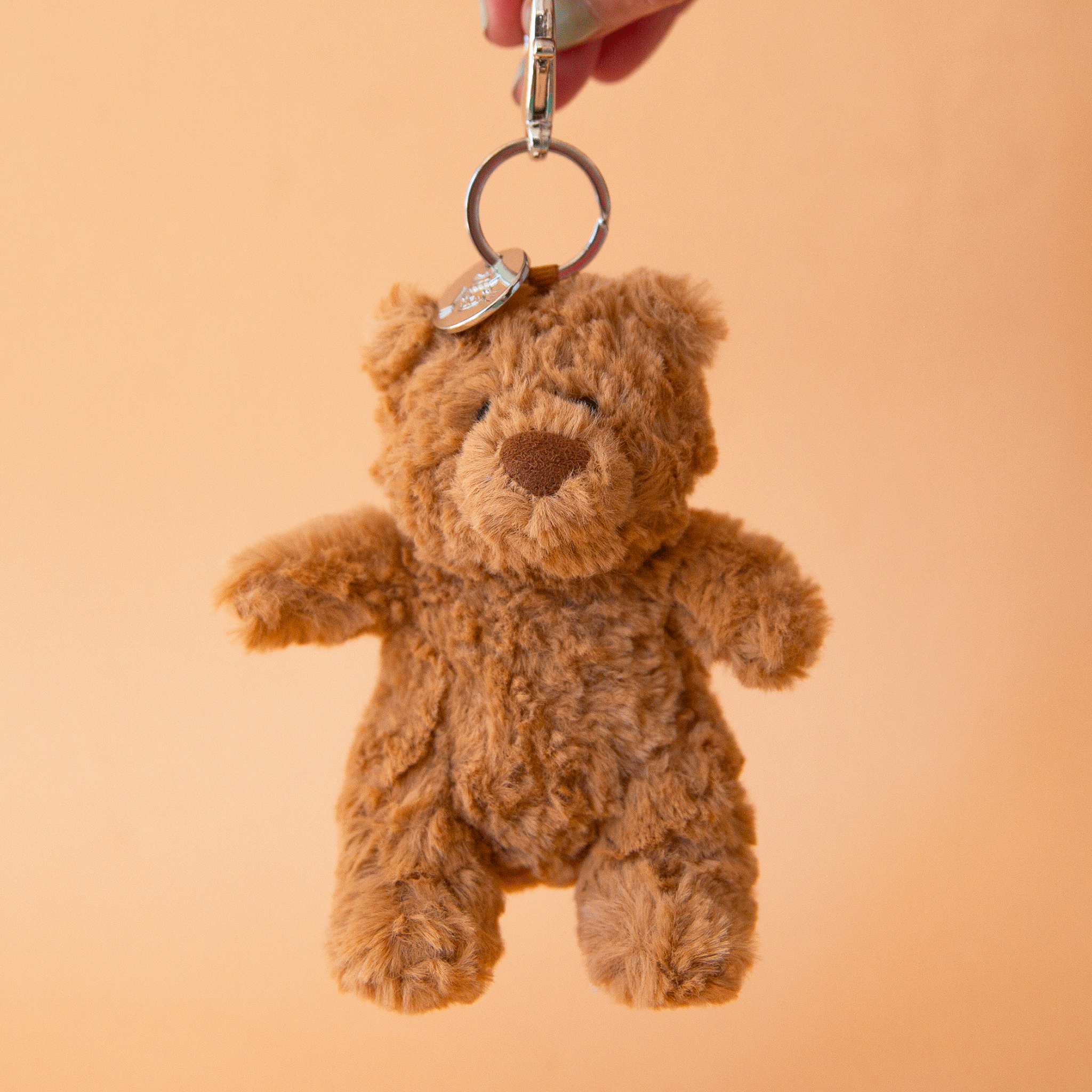 On a tan background is a brown bear stuffed toy on a silver loop and hook for adding to a bag or keychain. 
