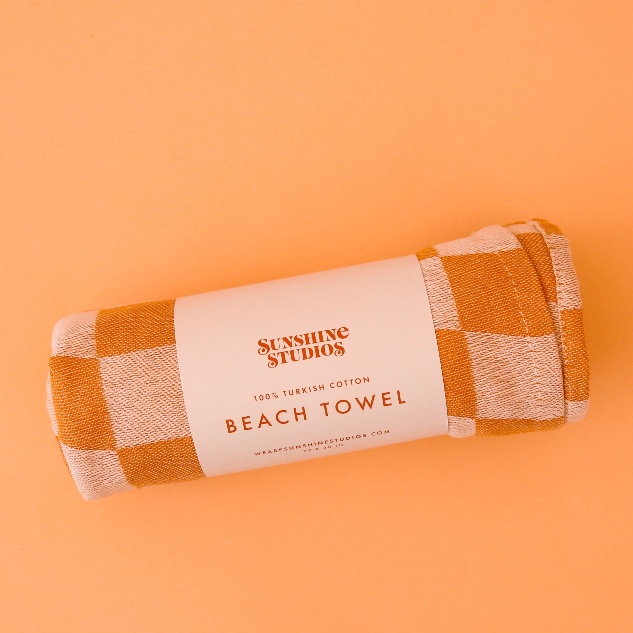 A rolled up beach towel with a orange checkered pattern laying on an opened up towel.