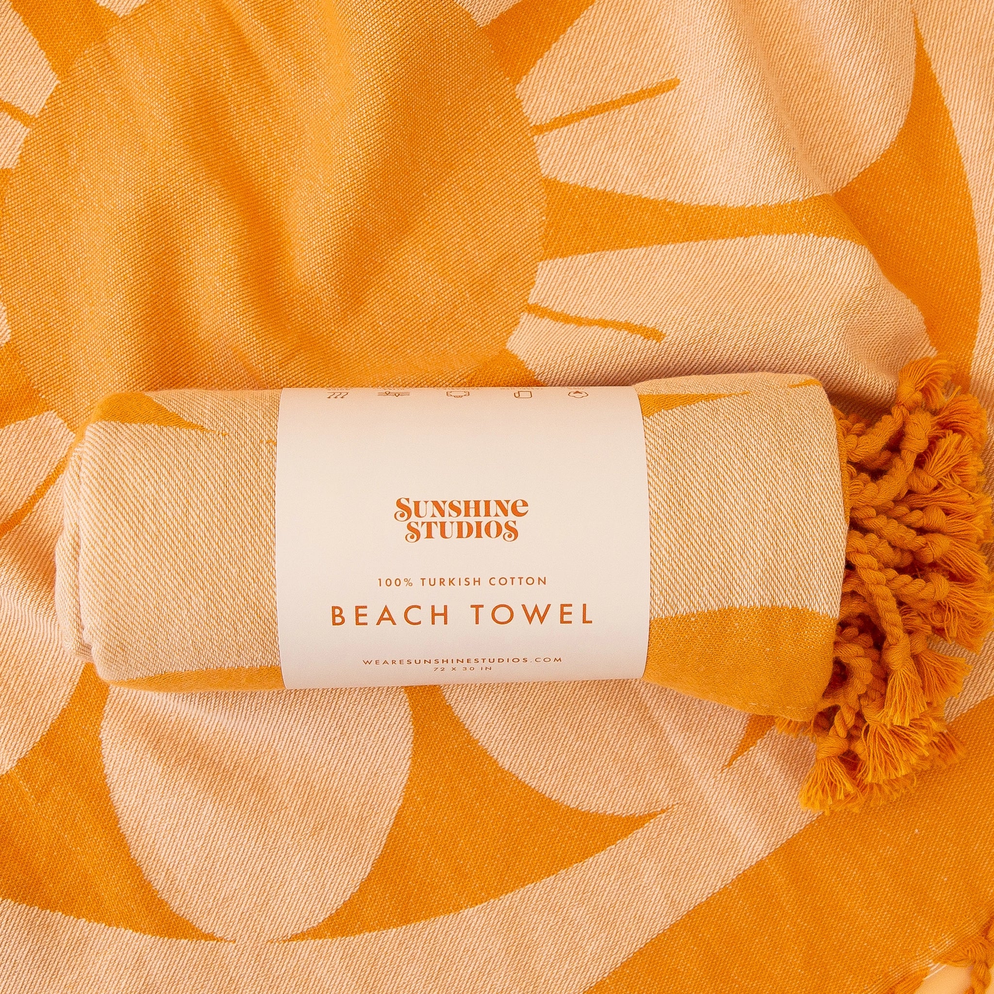 An orange beach towel rolled up with white label that reads, &quot;Sunshine Studios 100% Turkish Cotton Beach Towel&quot; along with a retro flower design and orange string tassels.