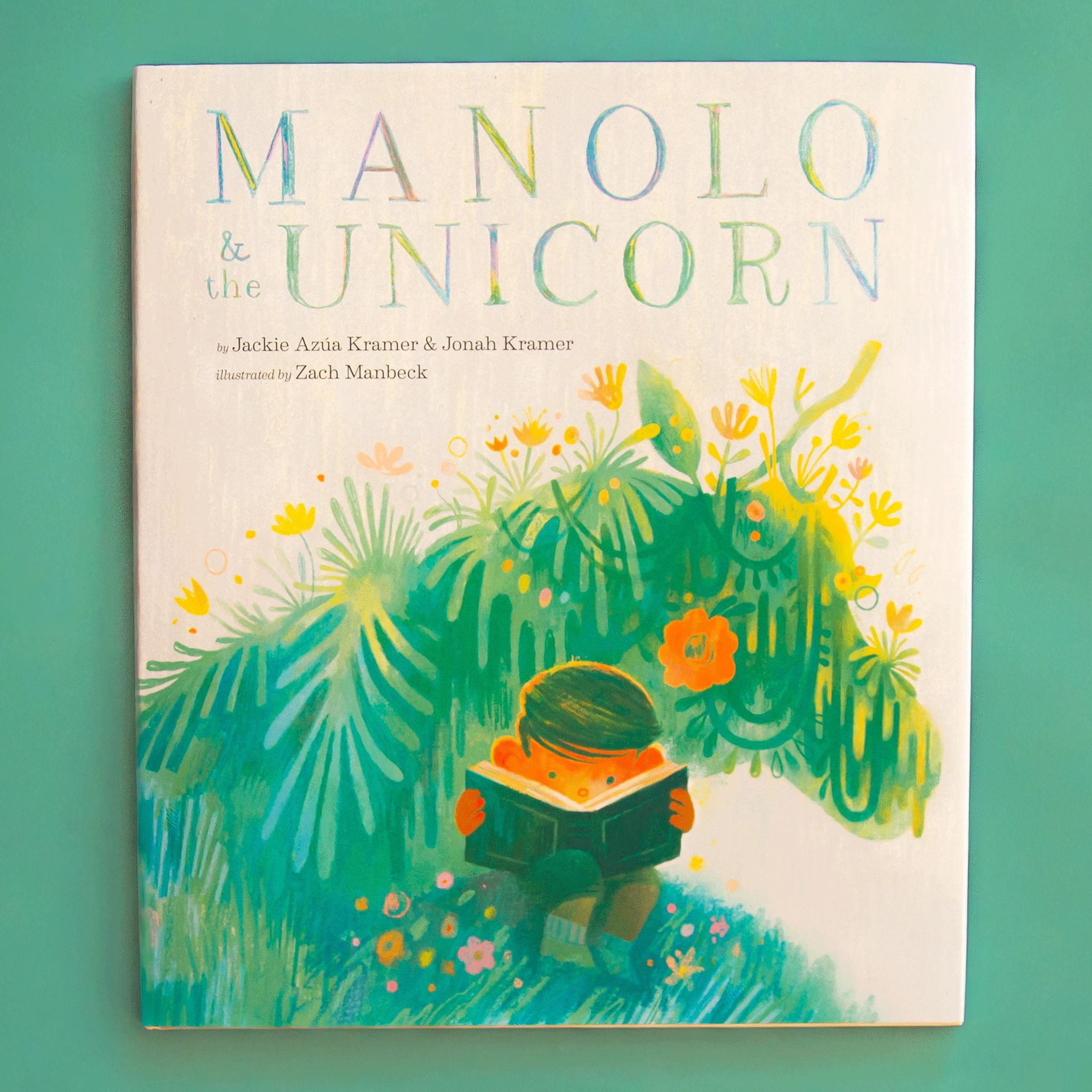 On a green background is a children&#39;s book with a title that reads, &quot;Manolo &amp; the Unicorn&quot; with a graphic of a child reading a book with a unicorn shaped greenery behind him. 