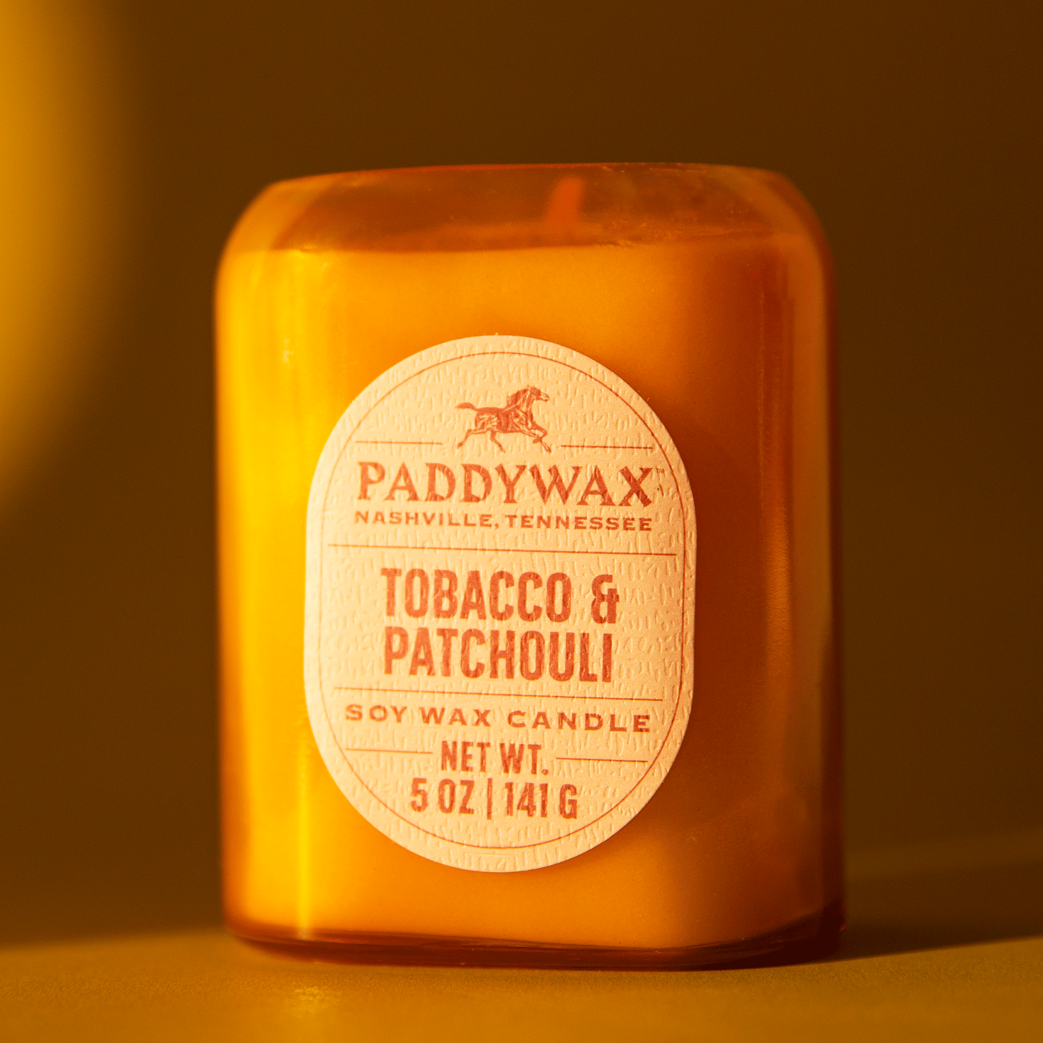 On a burnt orange background is a glass jar candle with an oval label in the center that reads, &quot;Paddywax Tobacco Patchouli&quot;. 