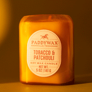 On a burnt orange background is a glass jar candle with an oval label in the center that reads, "Paddywax Tobacco Patchouli". 