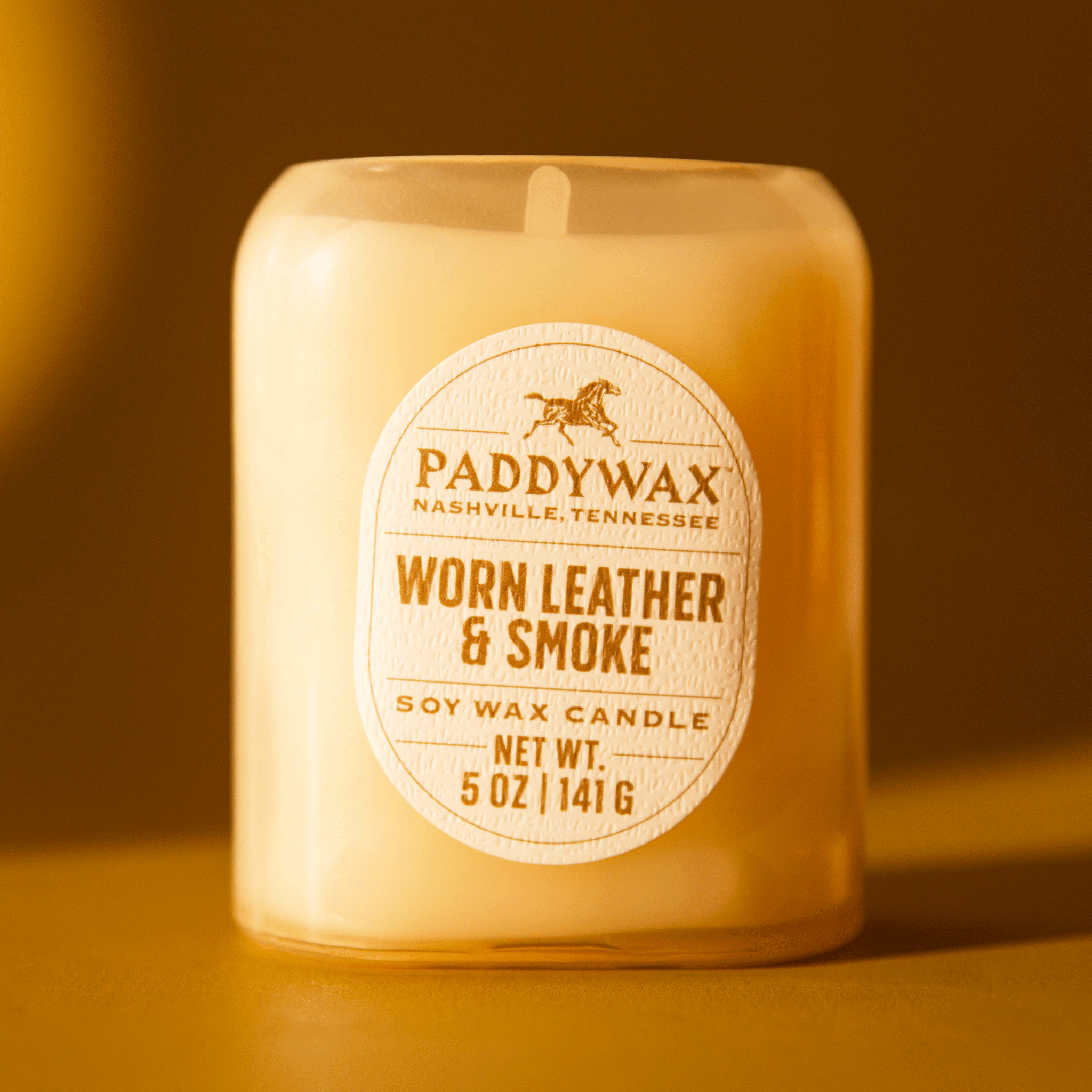 On an orange background is a neutral glass jar candle with an oval label that reads, &quot;Paddywax Worn Leather &amp; Smoke&quot;. 