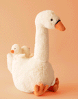On a orange background is a swan shaped stuffed animal toy with two baby swans on her back.