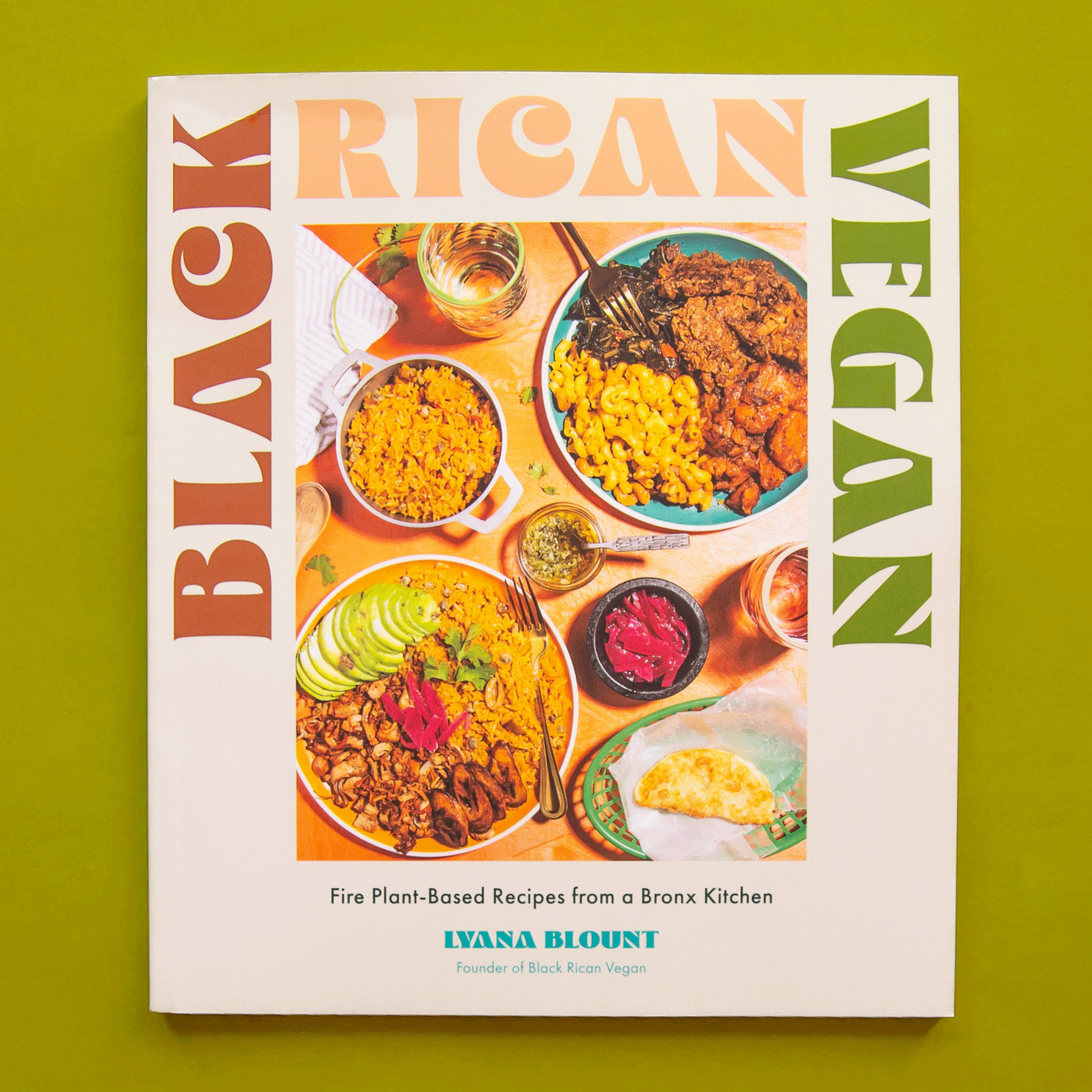 On a green background is a white cook book with a colorful table of food with a title that reads, &quot;Black Rican Vegan&quot;. 