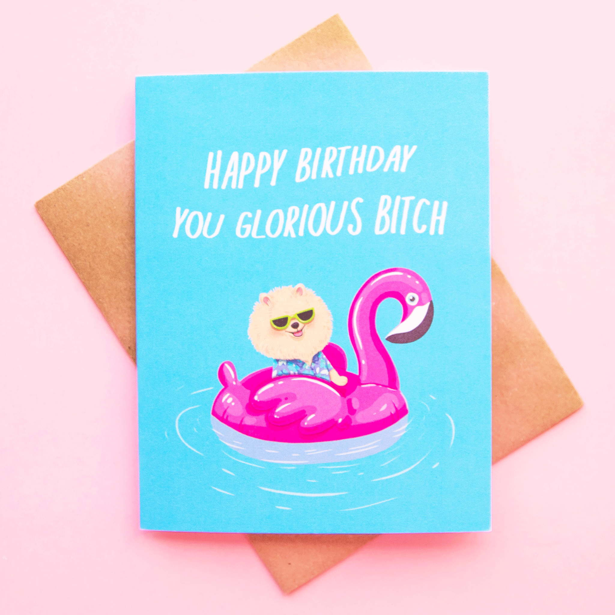 On a pink background is a bright blue card with an illustration of a Pomeranian in sunglasses on a pink pool float and white text that reads, &quot;Happy Birthday You Glorious Bitch&quot;.