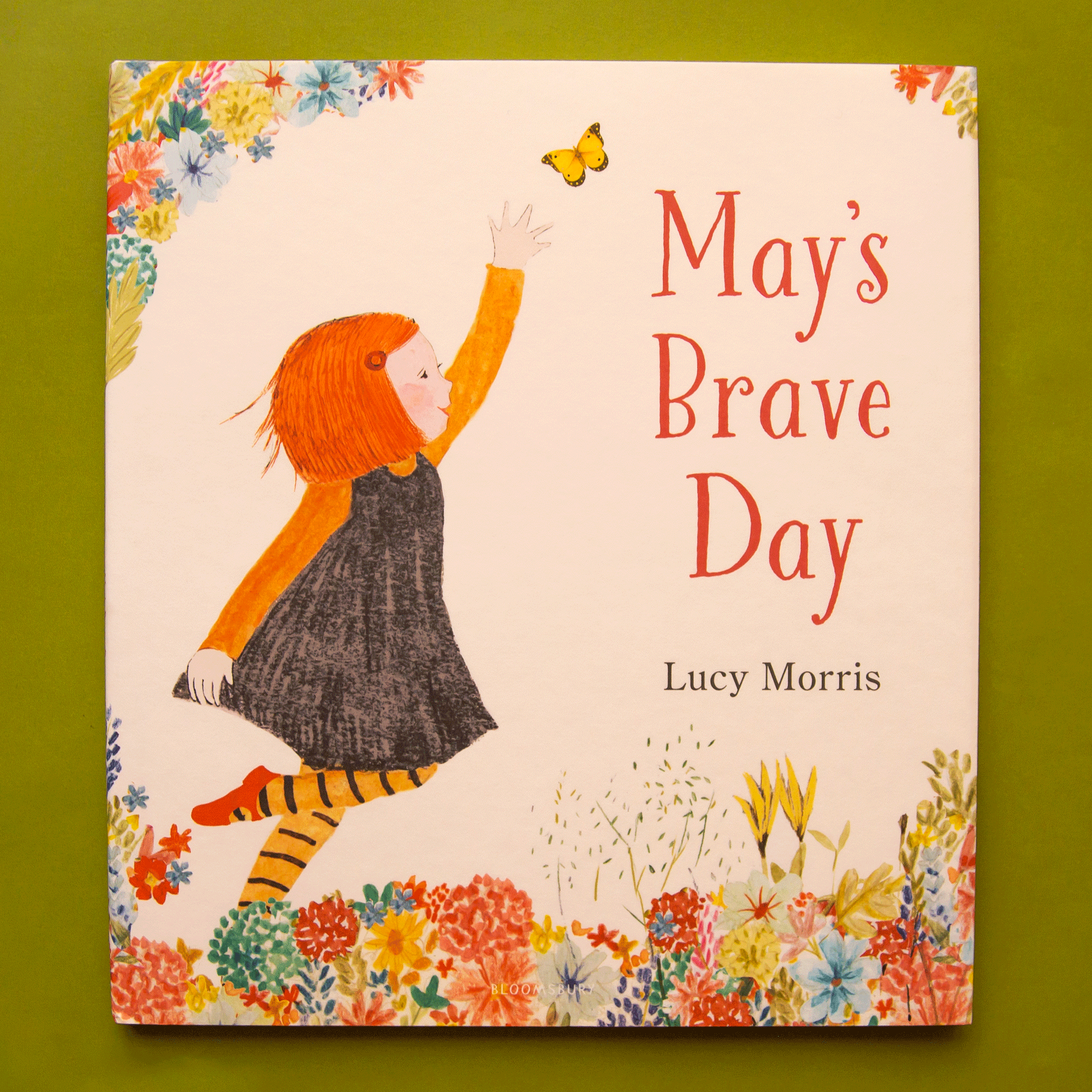 On a green background is a ivory book cover with an illustration of a little girl reaching for a yellow butterfly and text on the right side that reads, &quot;May&#39;s Brave Day&quot;. 
