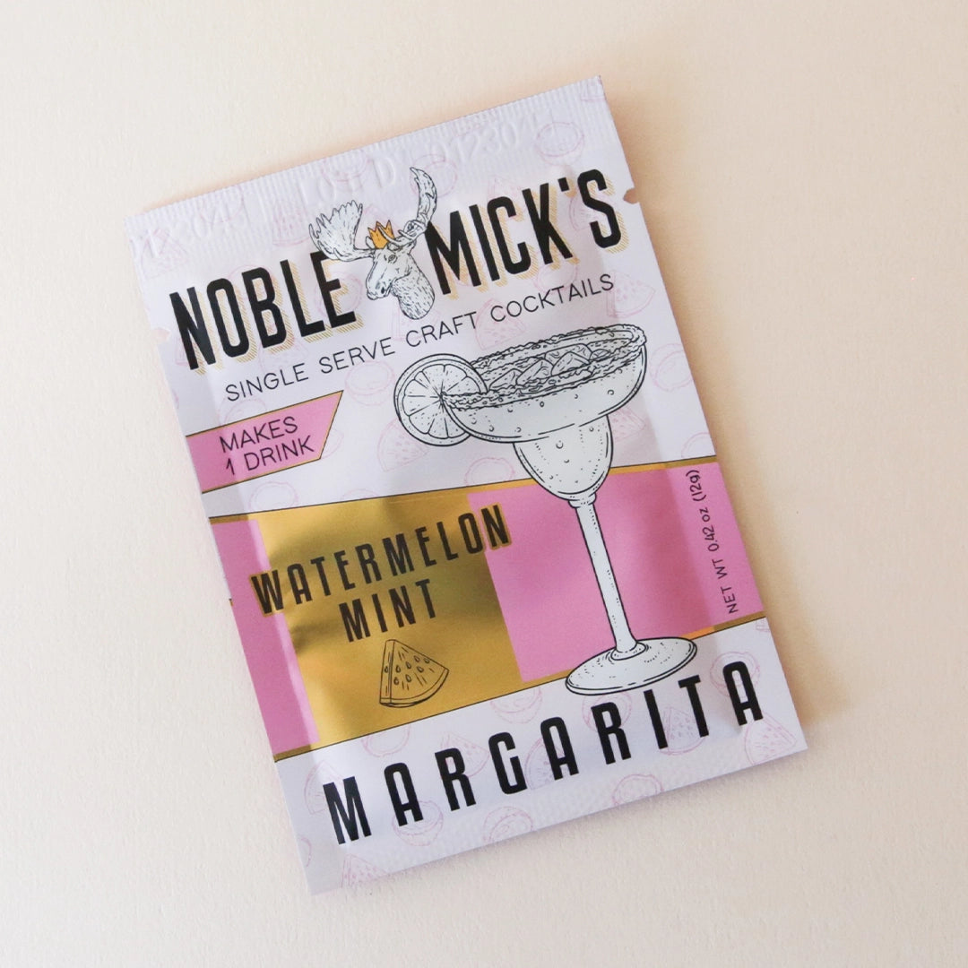 On a tan background is a packet of cocktail mix in a white and hot pink packaging that has an illustration of a margarita glass along with black text that reads, &quot;Noble Micks Single Serve Craft Cocktails, Watermelon Mint Margarita&quot;.