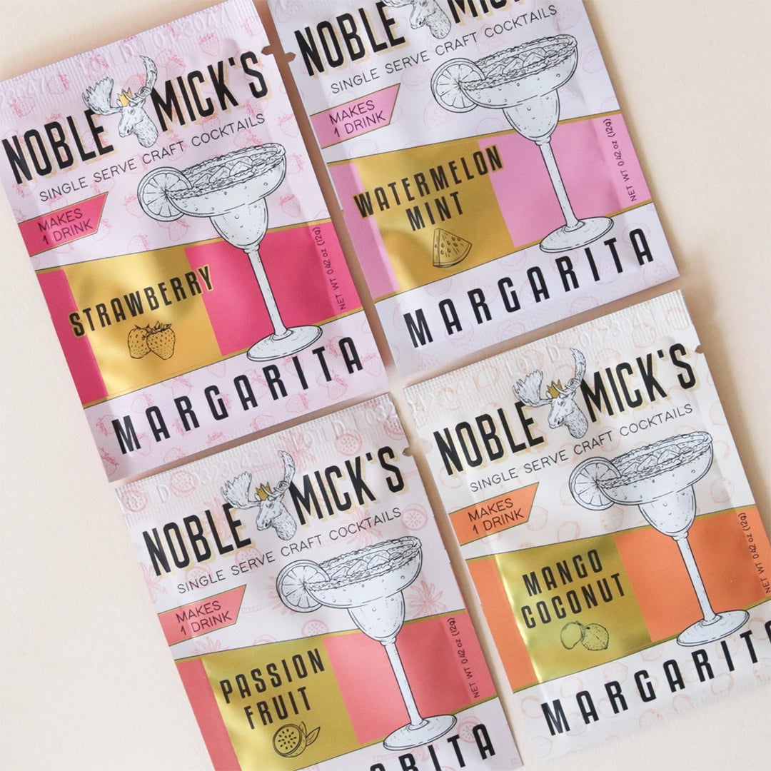 On a tan background is four different packets of Noble Mick&#39;s craft cocktail mixes with either pink, red or orange tones and imagery of a cocktail glass and a moss as well as text that reads, &quot;Noble Mick&#39;s Margarita&quot;.