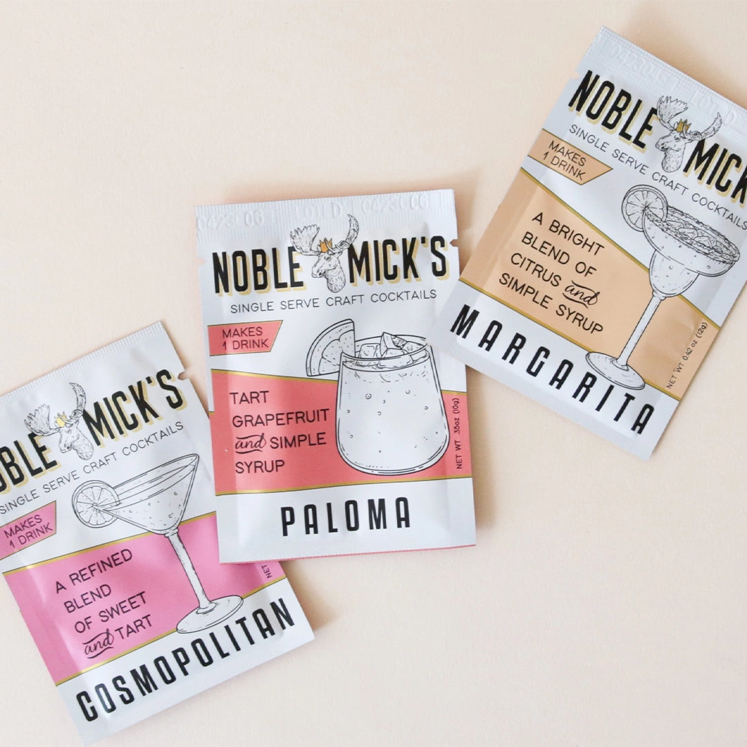 On a tan background is three packets of the Noble Mick&#39;s cocktail mixes in a variety of flavors.