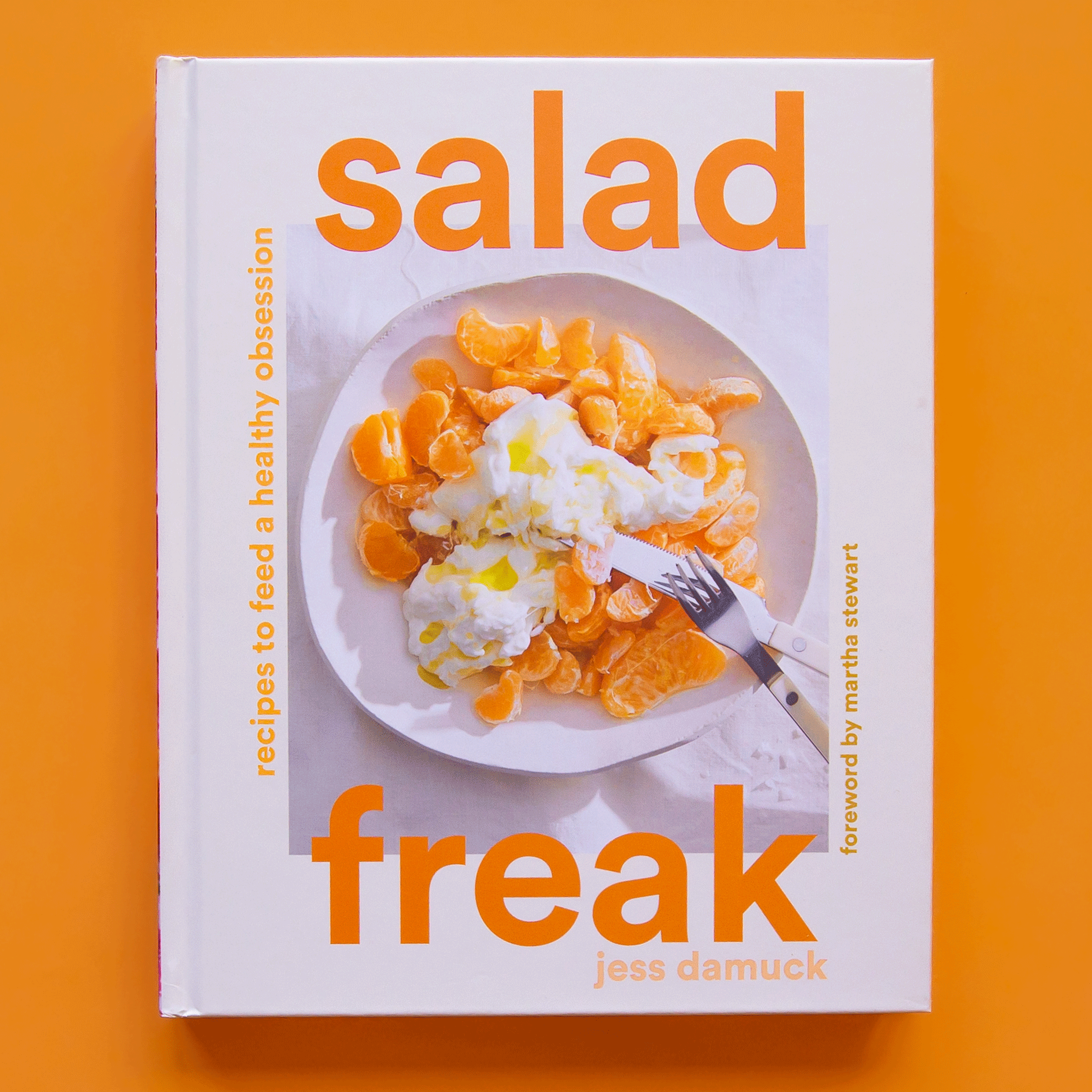 A white cookbook that has a bright orange text reading, "Salad Freak, Recipies to feed a healthy obsession". The cover also has a photo of a vibrant Clemintine orange salad.