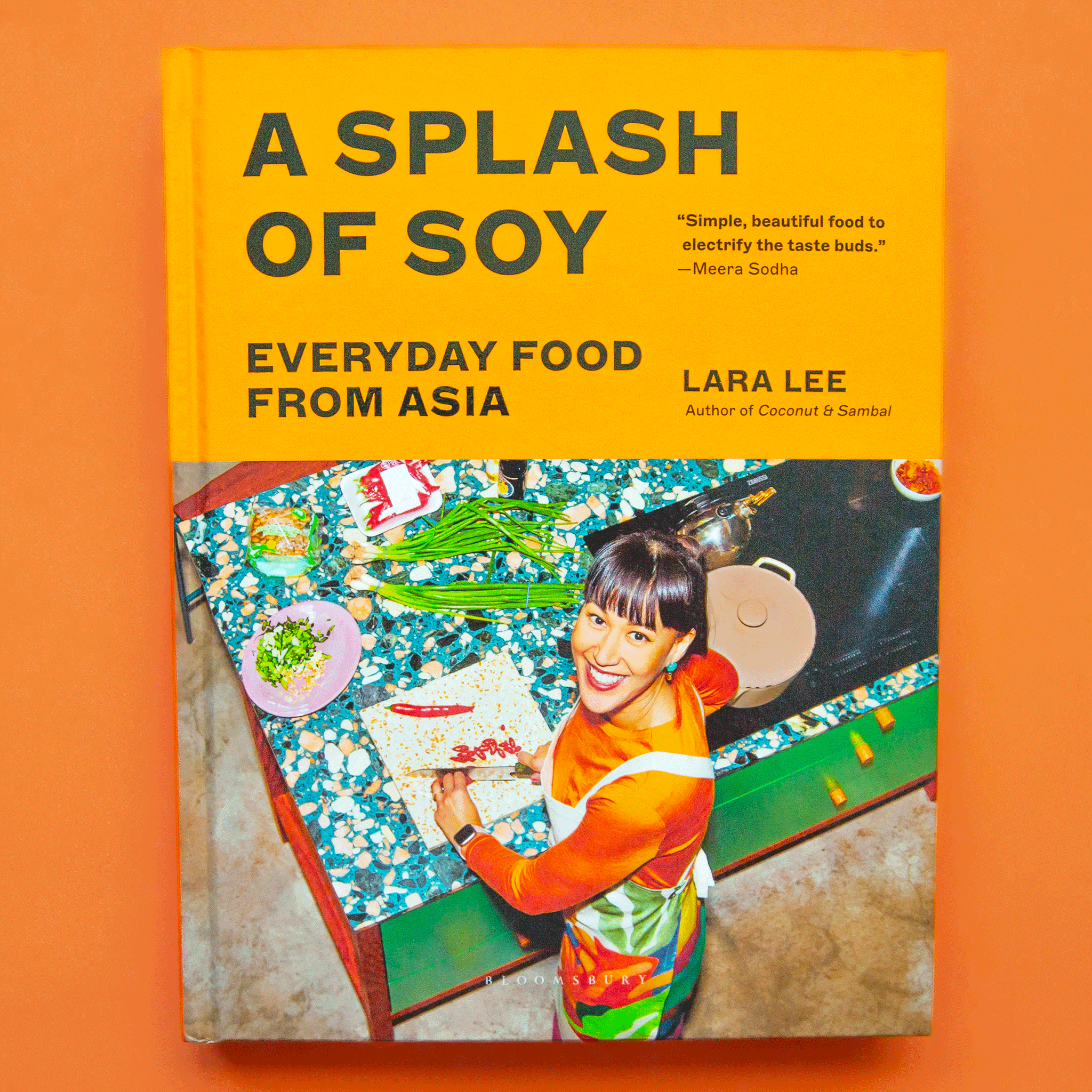 On an orange background is a yellow book with a photo of a woman cooking and the title that reads, &quot;A Splash Of Soy Everyday Food From Asia&quot;. 