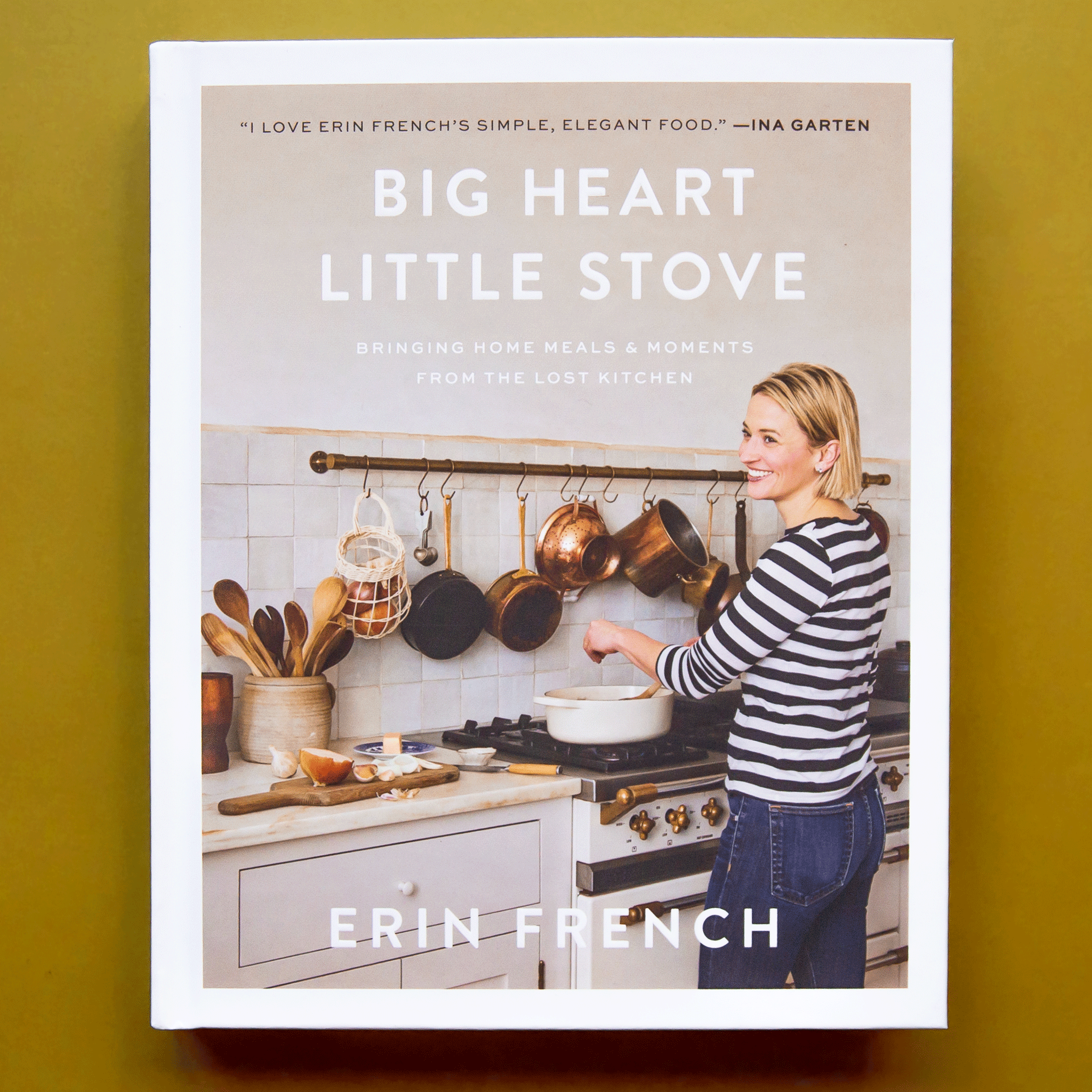 On a chartreuse background is a neutral colored bookcover with a photo of a woman cooking in front of the stove with the title toward the top that reads, &quot;Big Heart Little Stove Bringing Home Meals &amp; Moments From The Lost Kitchen&quot;. 