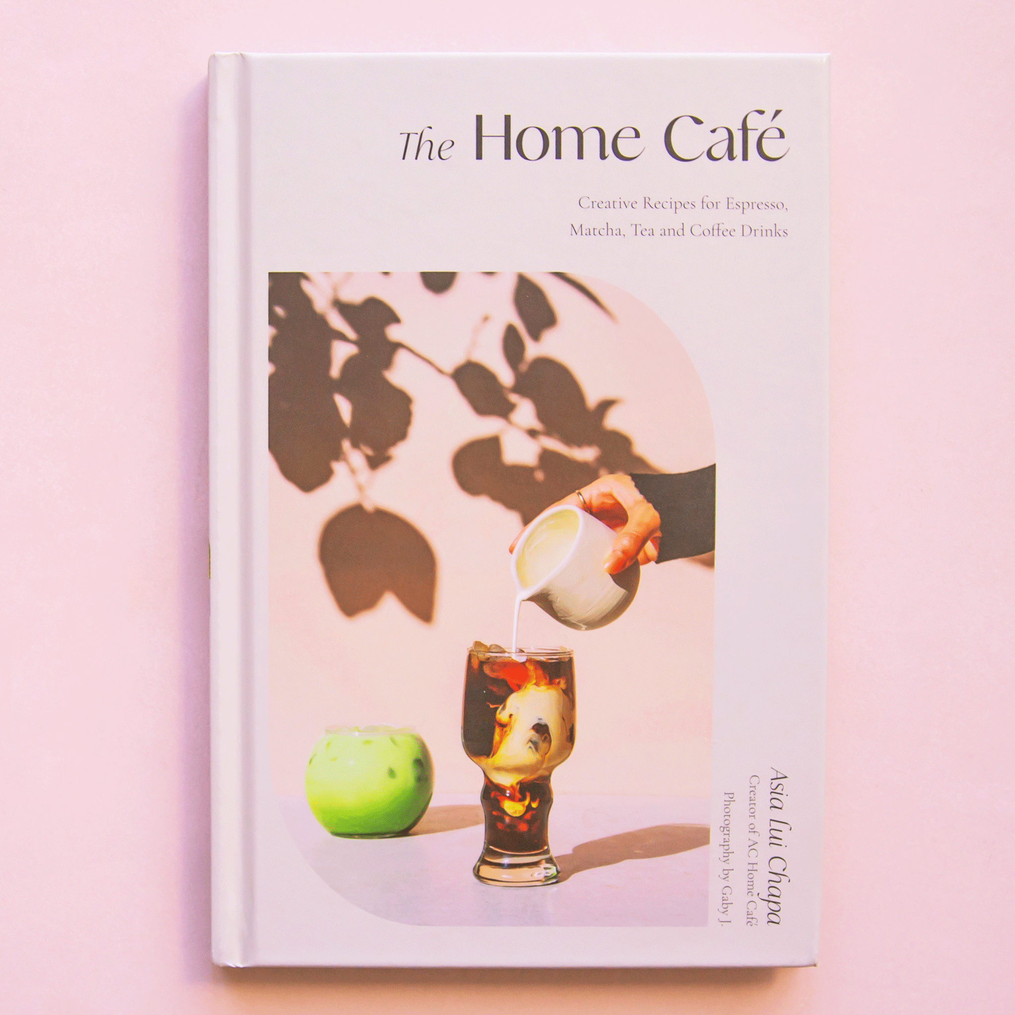 On a light pink background is an ivory book with a photograph of a coffee in a glass and cream being poured in along with the title of the book at the top that reads, &quot;The Home Café Creative Recipes for Espresso, Matcha, Tea and Coffee Drinks&quot;.