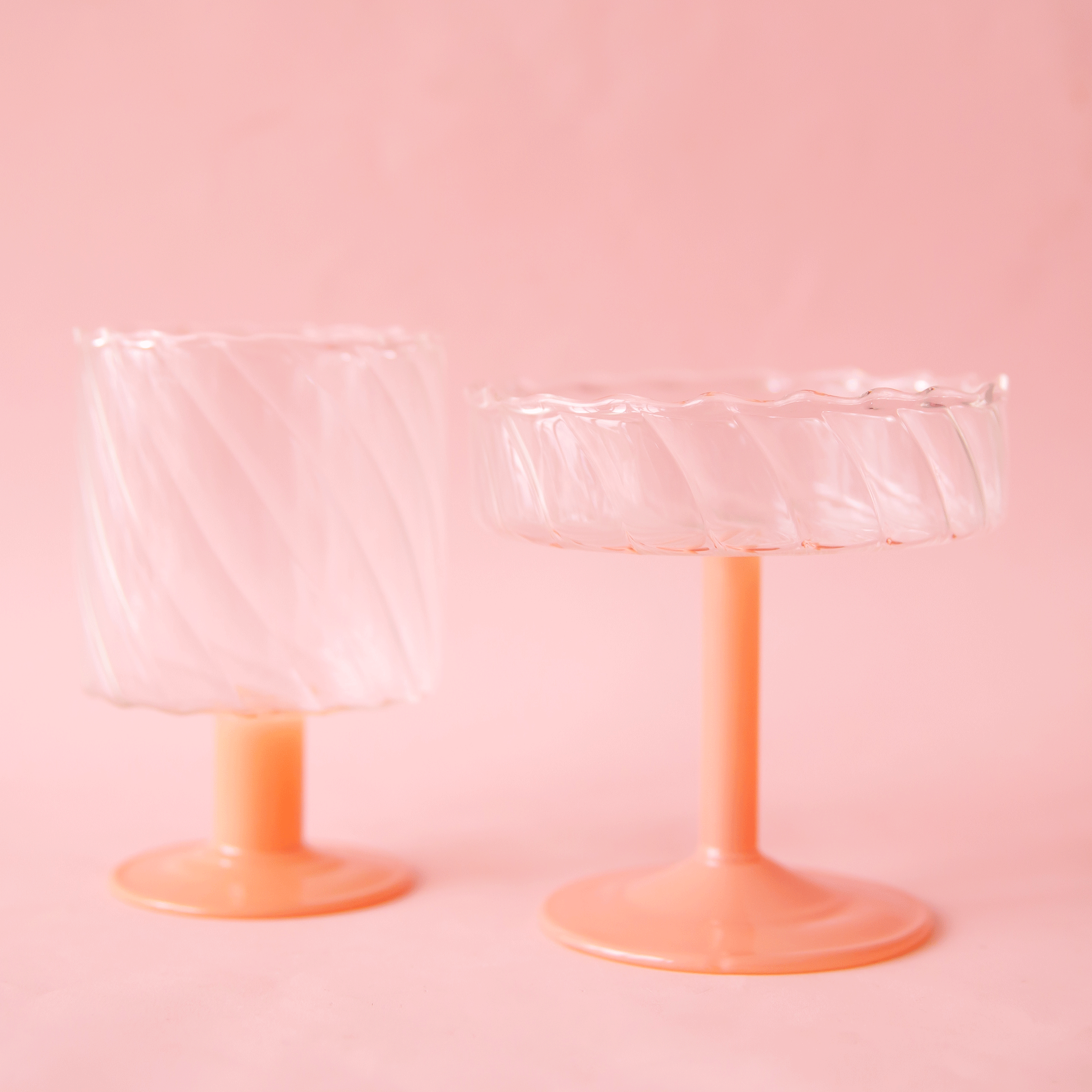 On a pink background is two wavy glass coupe with a pink stem. One glass is a coupe style, the other is a short wine glass style. 