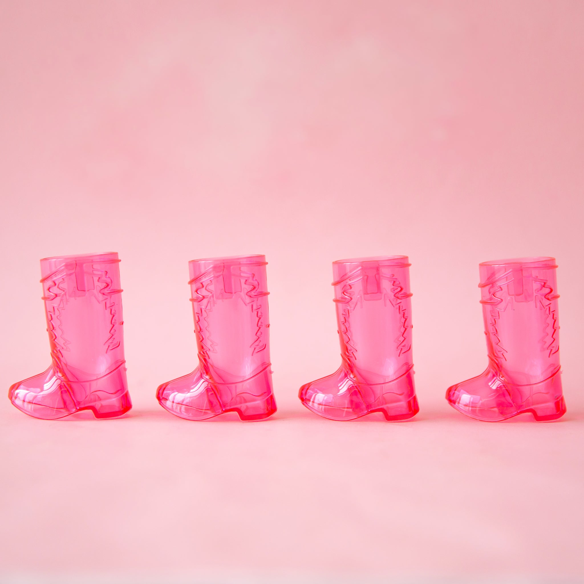 On a pink background is a pack of four plastic pink boot shaped shot glasses. 