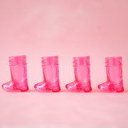 On a pink background is a pack of four plastic pink boot shaped shot glasses. 