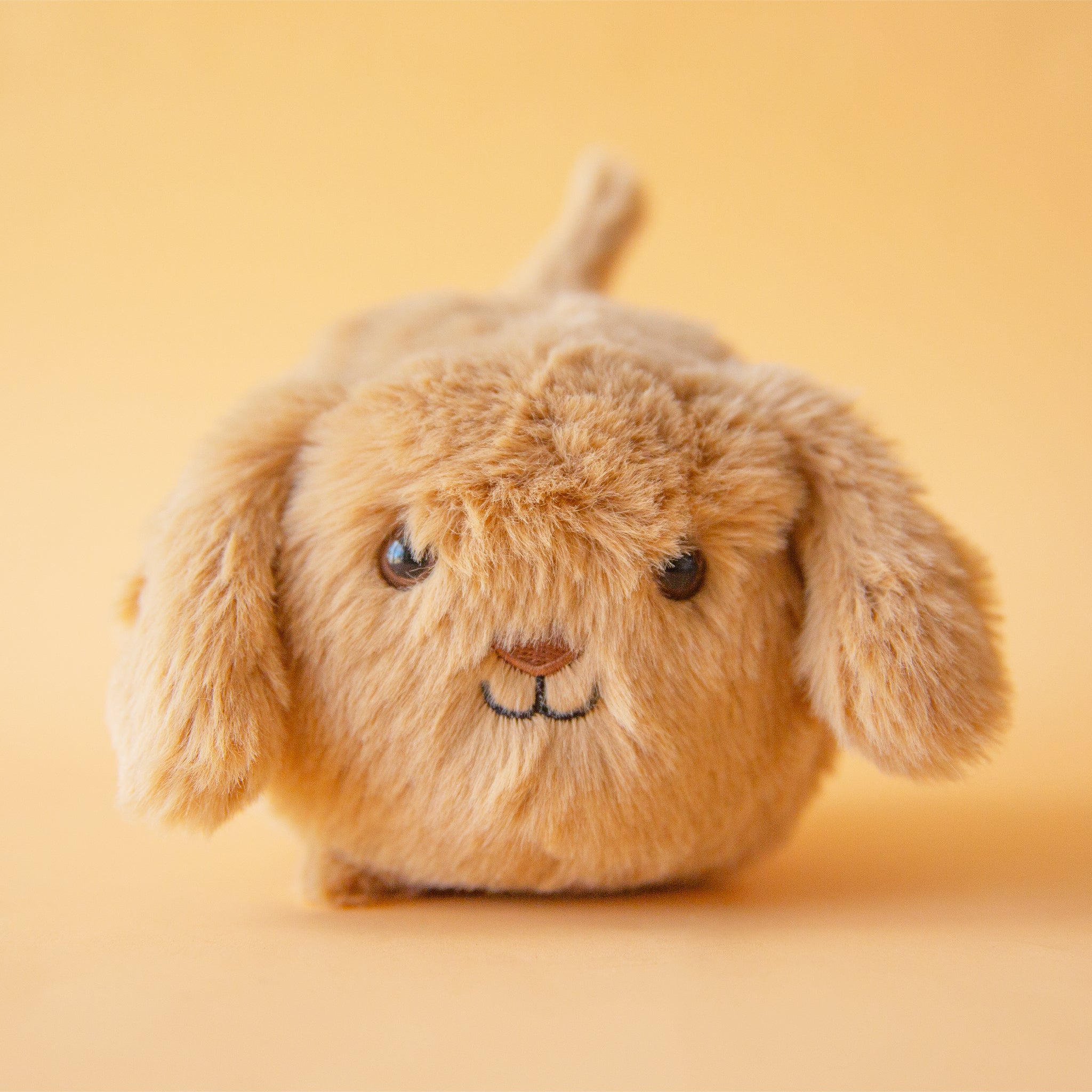 On a tan background is a tan puppy stuffed toy puppy with a round body and floppy ears.