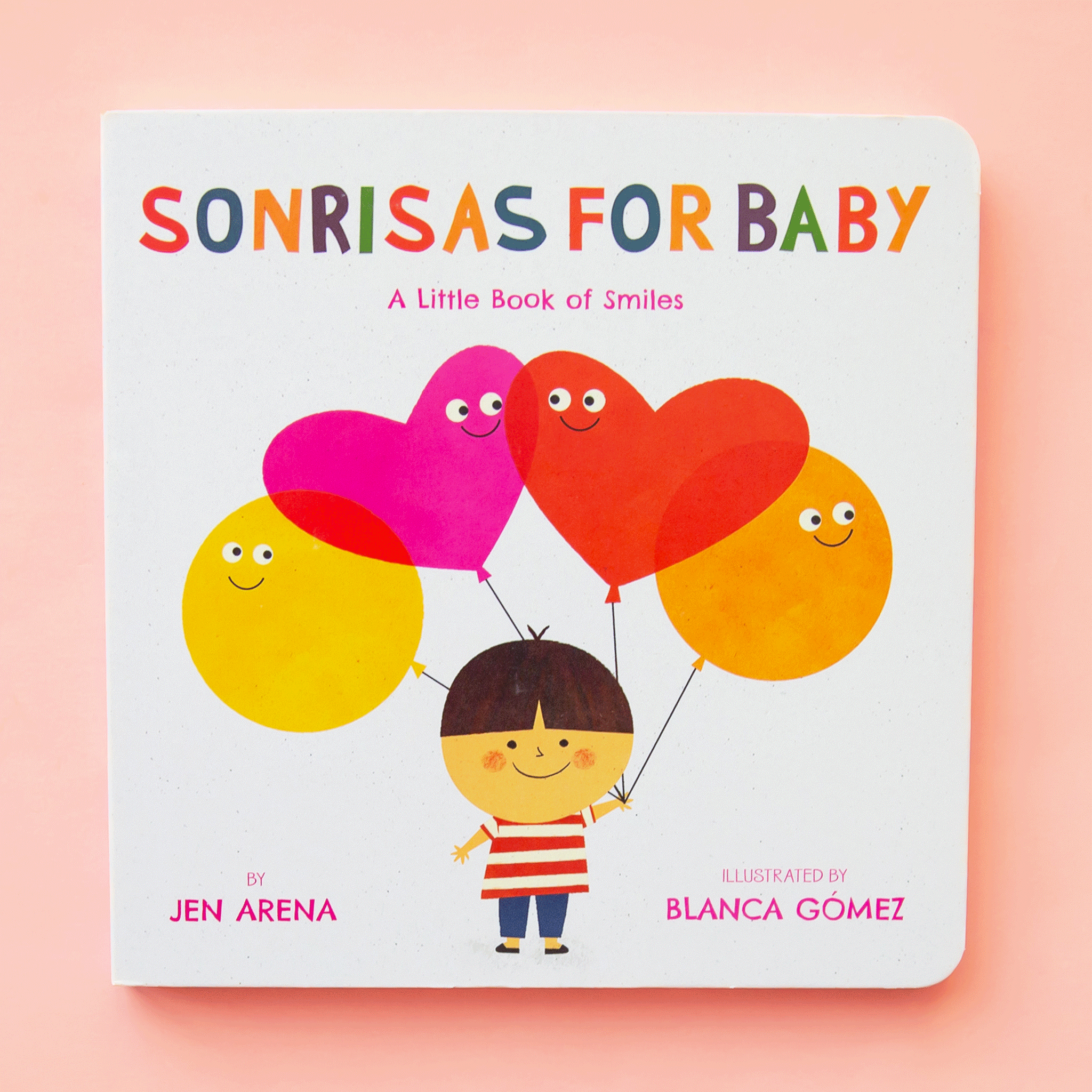 Cover of a white square book. In the middle is a drawing of a kid wearing a red and white striped t-shirt and blue pants. The kid has a large round head with brown hair and a smiley face. The child is holding four balloons. The left balloon is a yellow circle, the next is a pink heart, next is a red heart and the last balloon is an orange circle. All four balloons have a smiley face on them. At the top is rainbow text that reads ‘sonrisas for baby.’ Under is pink text that reads ‘a little book of smiles.'