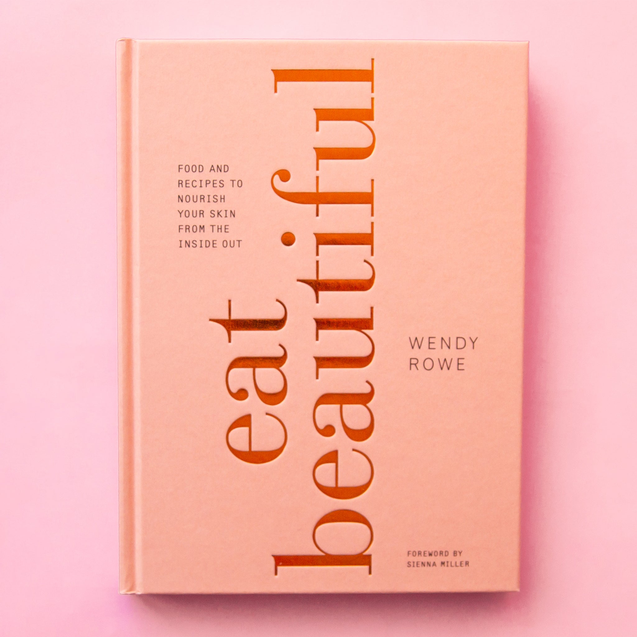 Cookbook with a soft pink hardcover that reads &#39;eat beautifully&#39; in large gold foil lettering facing perpendicular to the cover. In smaller lettering reads &#39;food and recipes to nourish your skin from the inside out&#39; in smaller lettering.
