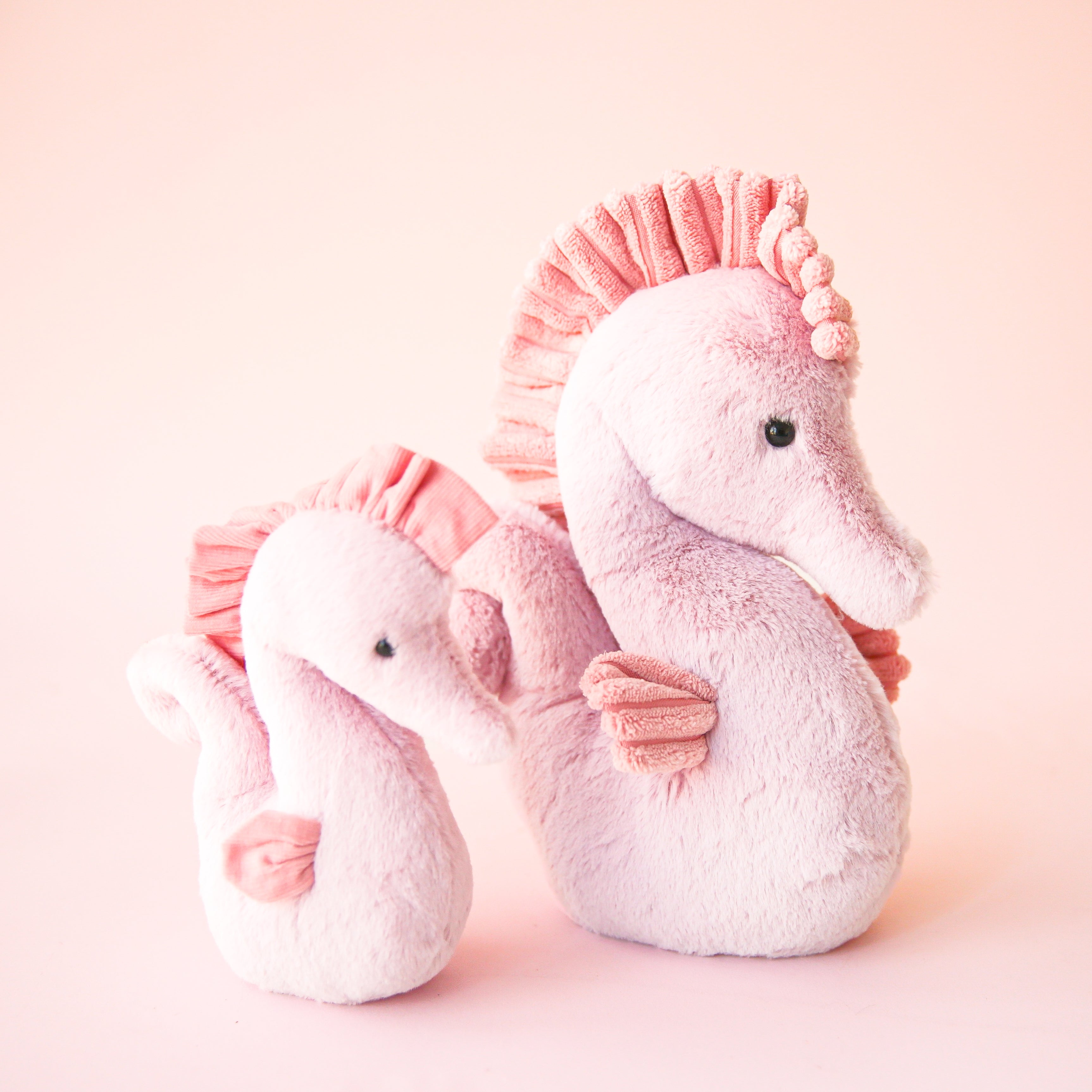 On a peachy background is two different sized pink seahorse shaped stuffed animals. 
