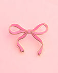 A pink enamel bow pin sits on top of a pink background. There is a gold brass border around the enamel lapel pin.