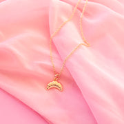 gold croissant shaped charm hangs on a gold necklace chain. the backrgound of the image is a soft pink chiffon fabric.