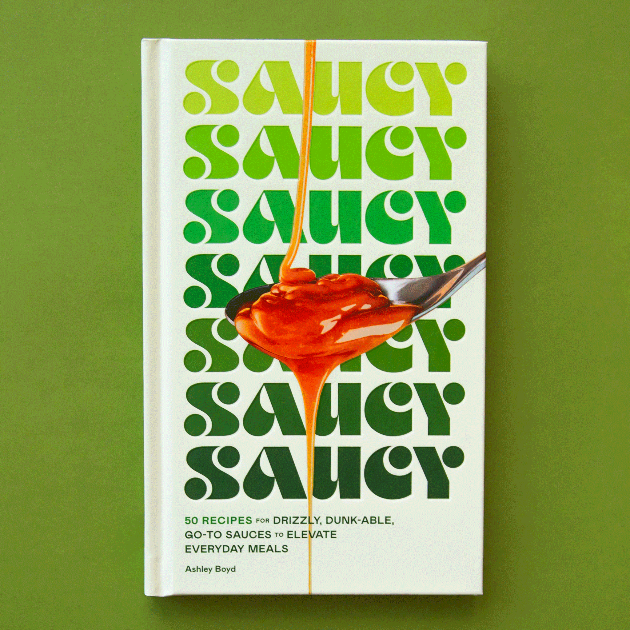On a green background is a white book with green repeated text that reads, &quot;saucy&quot; and a spoon graphic with a sauce drizzling off it. 