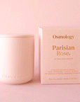On a pink background is a pink clay jarred candle with a lid and the box that it comes in on the side that reads, "Parisian Rose for mind body and soul.".