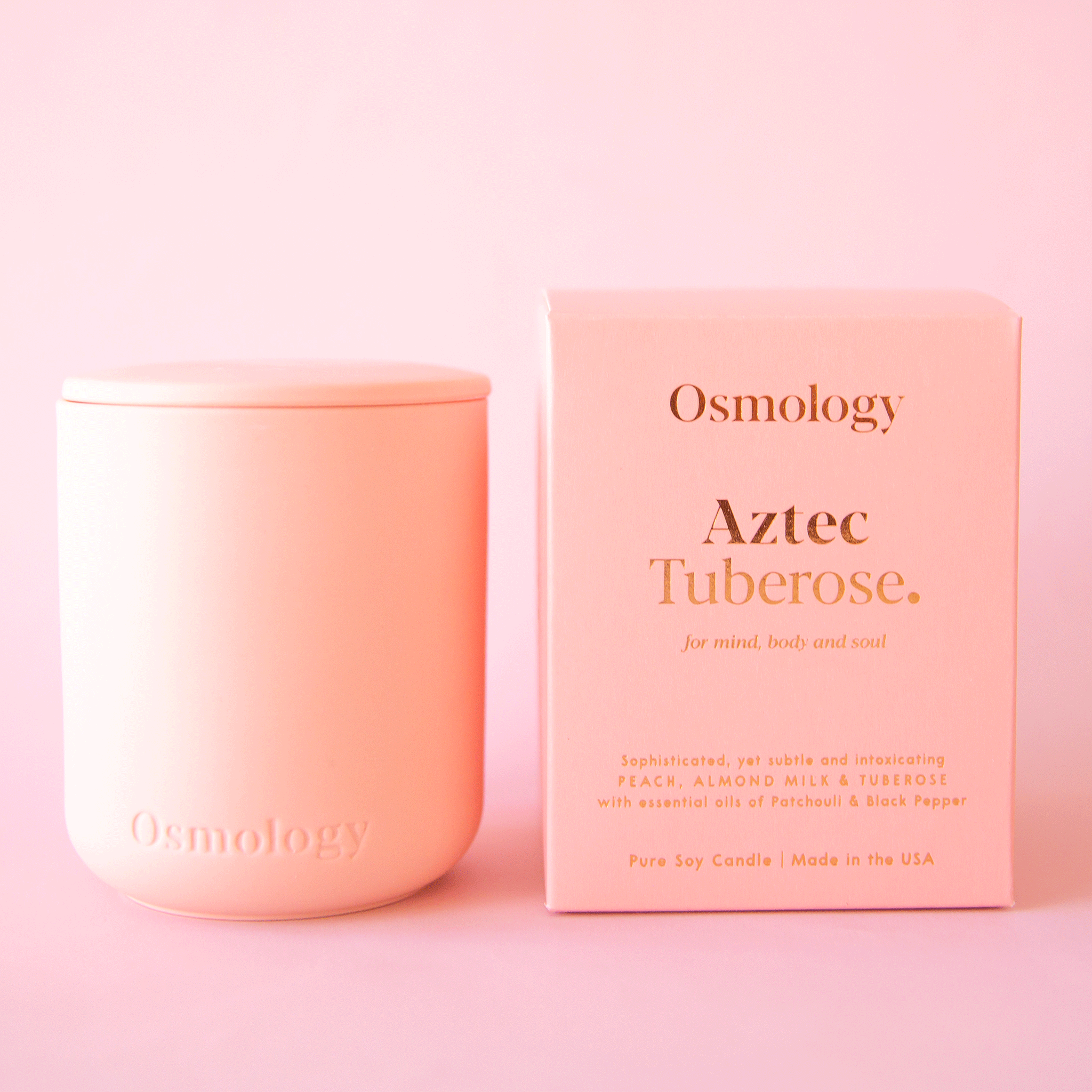On a pink background is a peach clay candle with a lid sitting next to the peachy box that it comes in. The front of the box reads, &quot;Osmology Aztec Tuberose.&quot;.