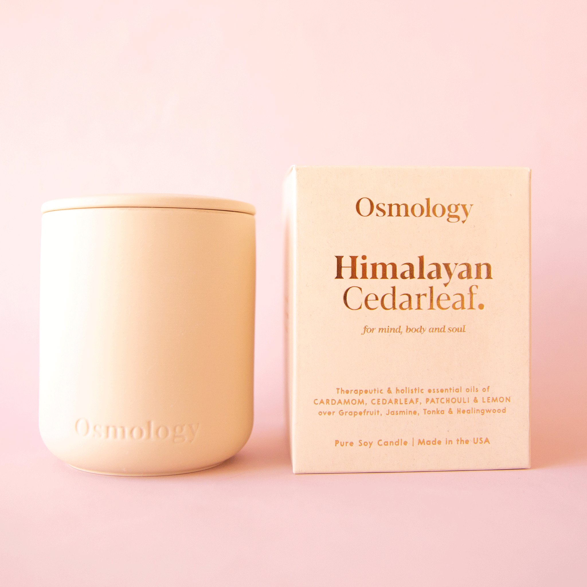 On a pink background is a peachy, tan clay candle with a lid and packaged in a coordinating box that reads, &quot;Osmology Himalayan Cedarleaf.&quot;.