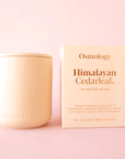 On a pink background is a peachy, tan clay candle with a lid and packaged in a coordinating box that reads, "Osmology Himalayan Cedarleaf.".