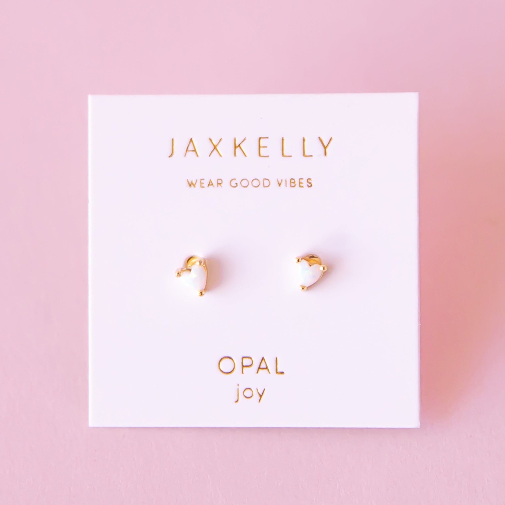 On a pink background is a pair of tiny heart shaped stud earrings with a white opal center.