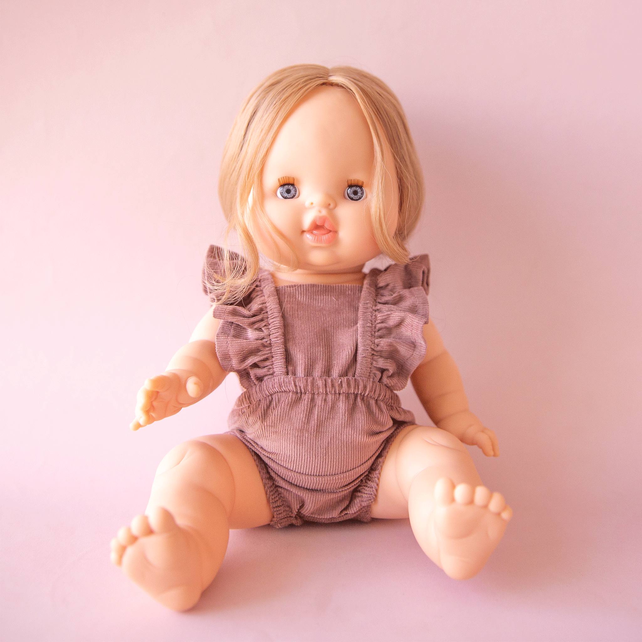 On a pink background is a baby doll with blonde braided hair and blue eyes wearing a pink romper not included with purchase. 
