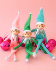 Three holiday elves sit on a pink background on top of hot pink glass ball ornaments. The elves are in a pink, green and turquoise elf outfit with tall hats and tinsel neck details.