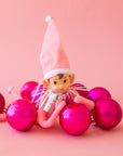 An adorable pink elf wearing a pink tall hat with silver tinsel around it's neck rests on hot pink christmas round ball ornaments.
