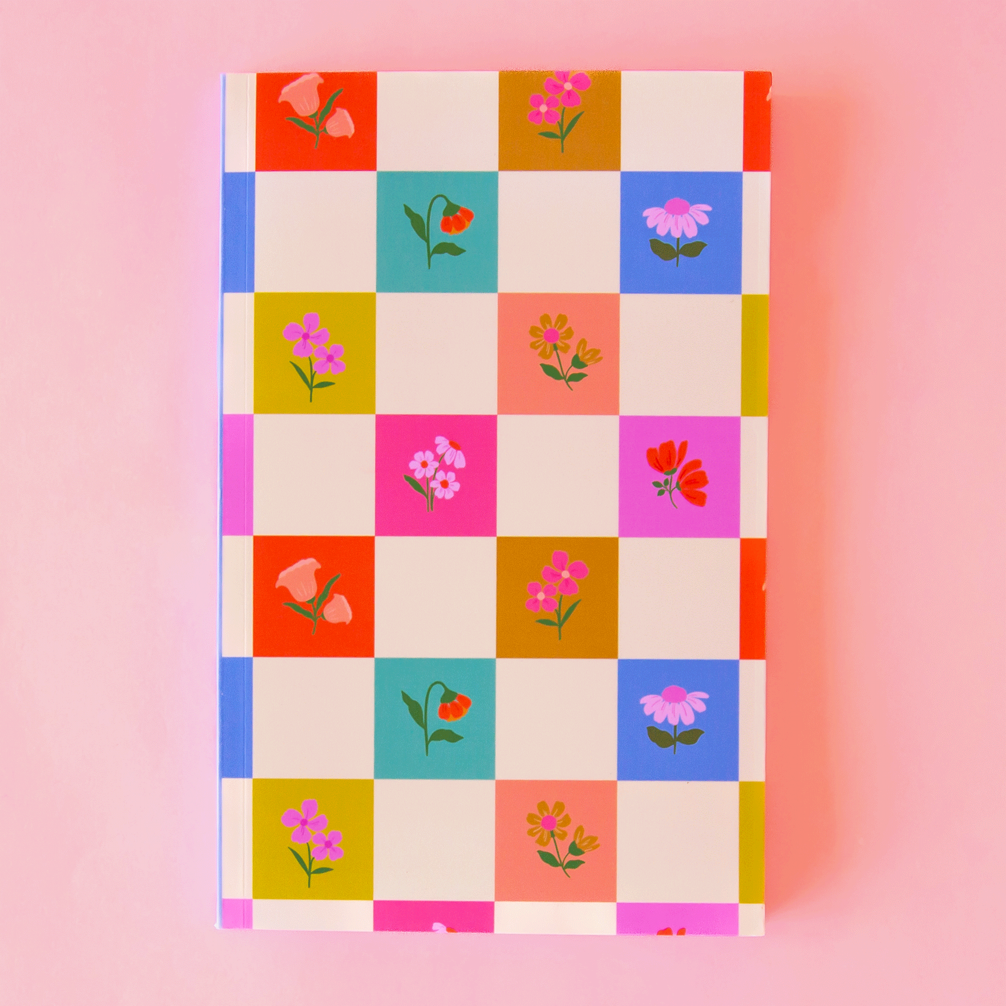 On a pink background is a multi colored checkered notebook with flower graphics in each of the colored squares. 
