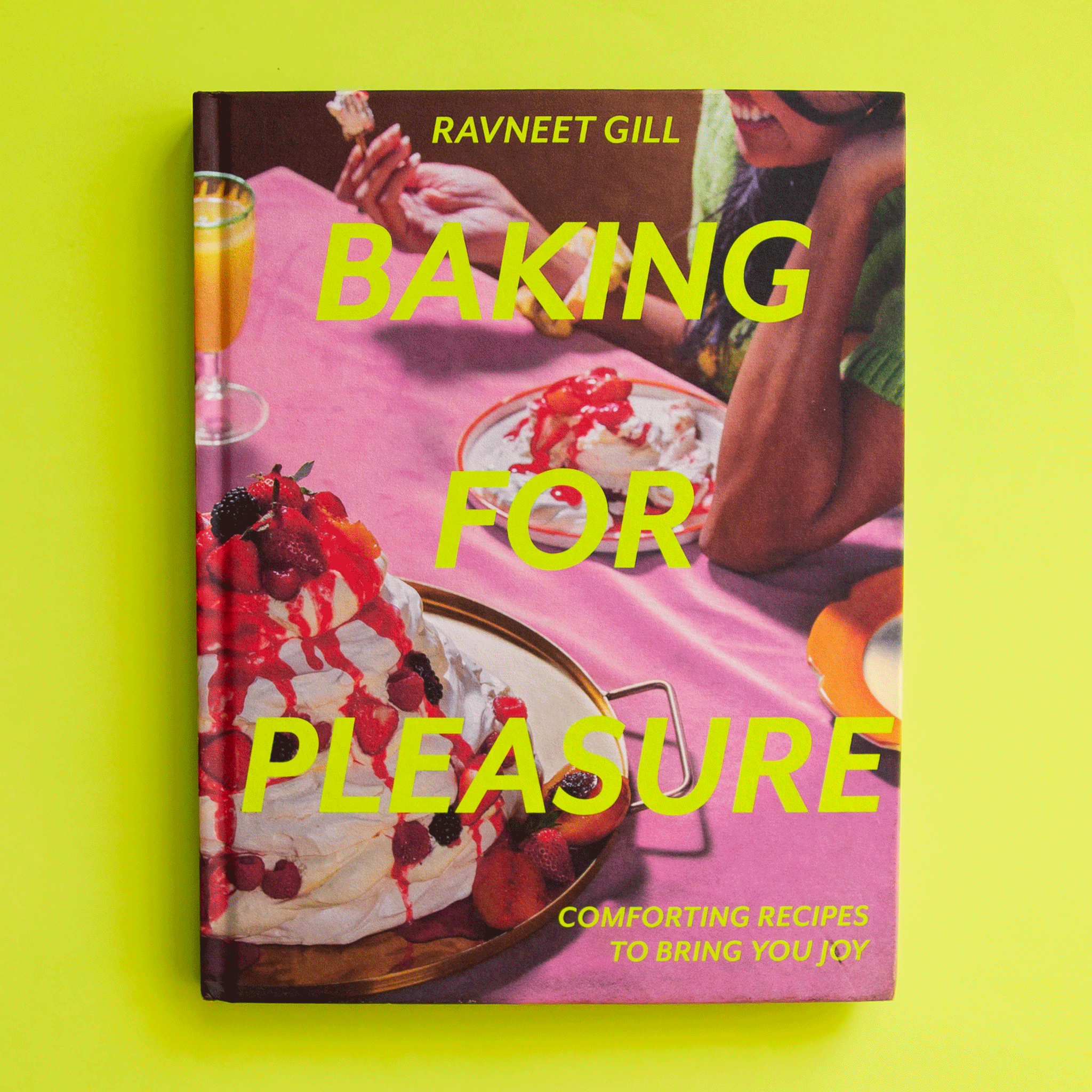 On a green background is a hot pink book cover with desserts on the front and yellow text that reads, &quot;Baking For Pleasure&quot;.