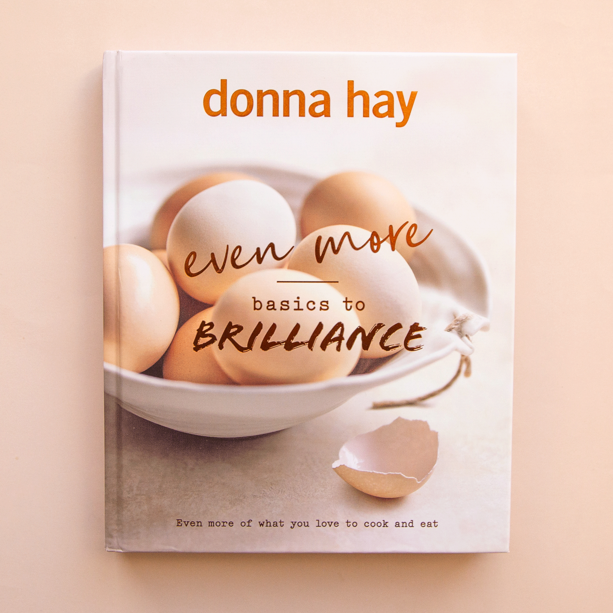 On a tan background is a neutral book cover with a photo of a bowl of eggs and the title that reads, "even more basics to Brilliance". 