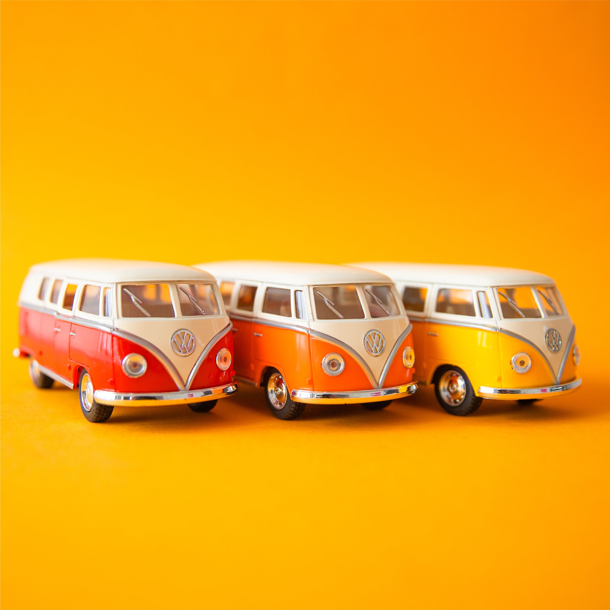 On a yellow background is three volkswagen bus toys in a red shade, an orange and a yellow. Each color sold separately.