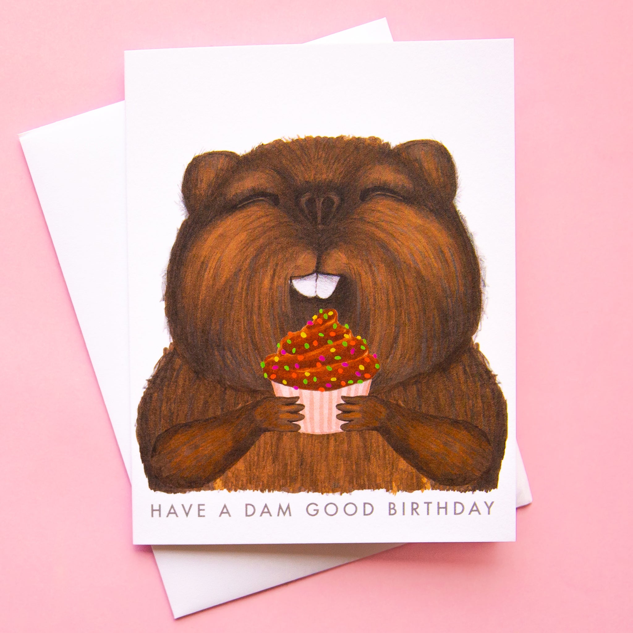On a pink background is a white card with an illustration of a brown beaver eating a cupcake and text below that reads, "Have A Dam Good Birthday". 