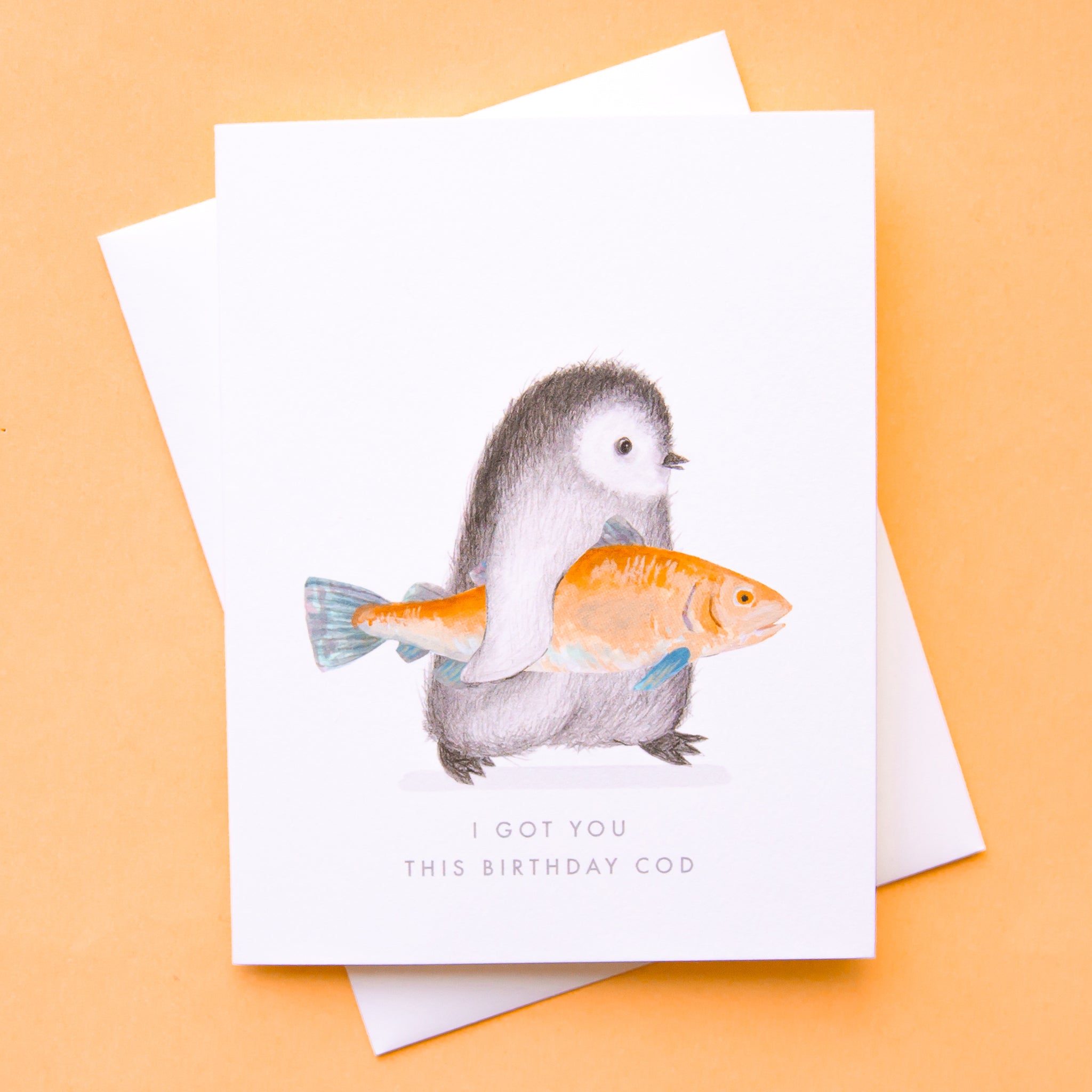 On a peach background is a white card and envelope that has a illustration of a penguin penguin holding an orange fish and text below that reads, &quot;I Got You This Birthday Cod&quot;. 