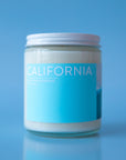 On a blue background is a clear glass jar candle with a light blue rectangular label across the front the reads, "California" in white letters and a white screw on lid.