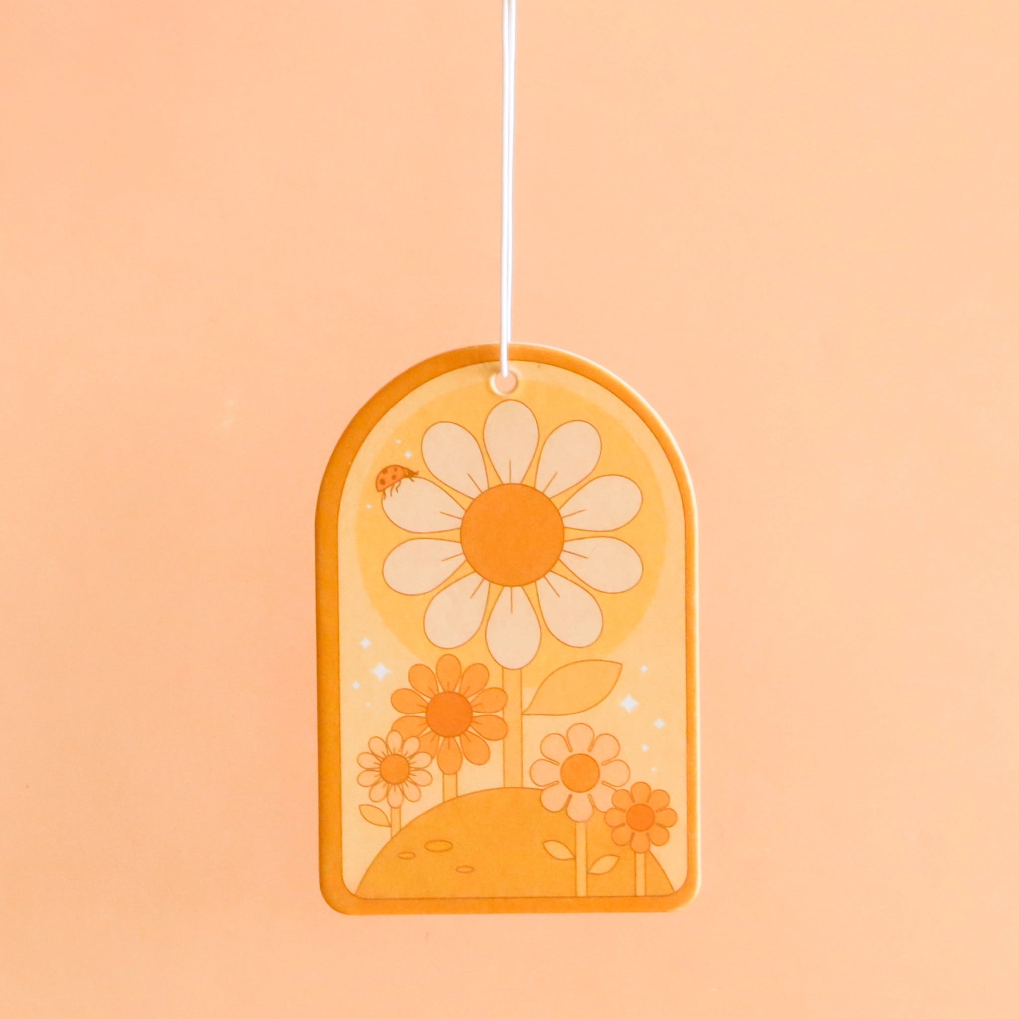 On a peachy background is an arched shaped air freshener with daisy designs and a white elastic loop for hanging.