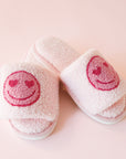 Pink fuzzy slide slippers with a darker pink heart eyes smiley face graphic on the front of each slipper.