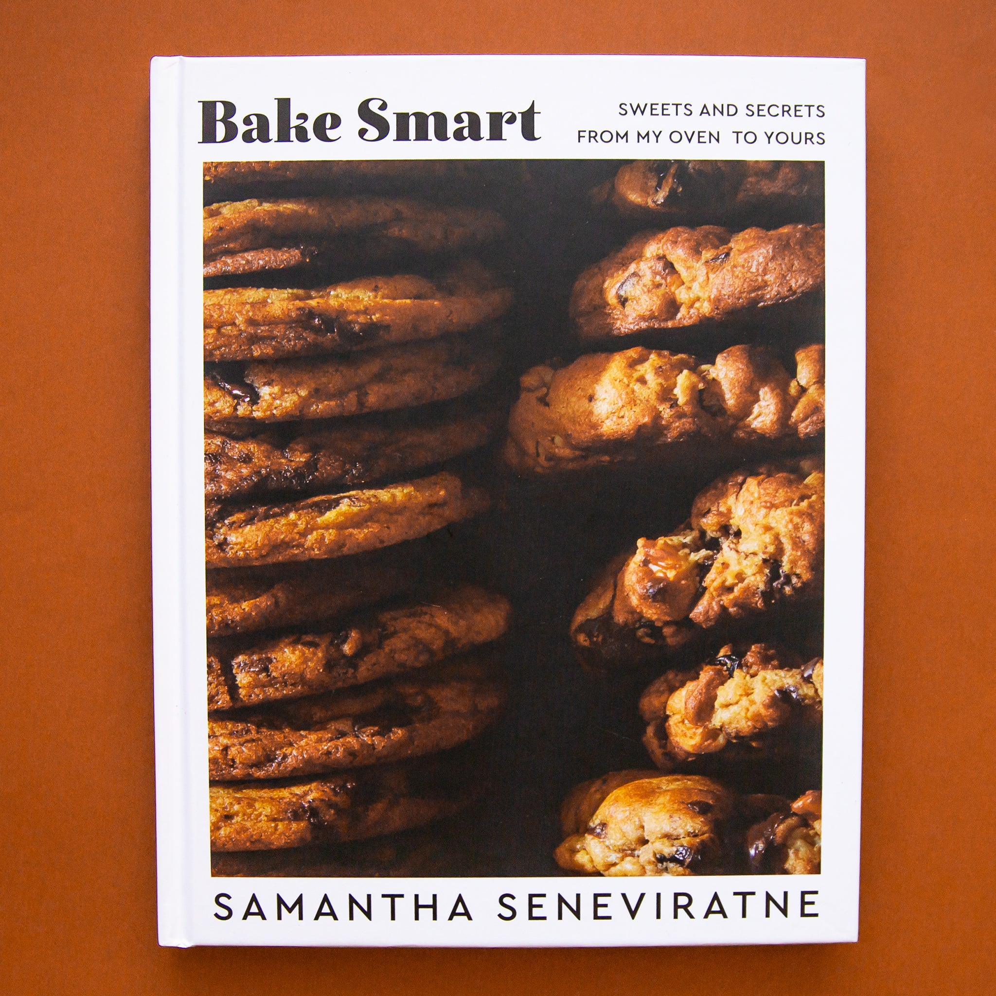 On a burnt orange background is a white book cover with a photo of cookies and the title on the top that reads, &quot;Bake Smart Sweets And Secrets From My Oven To Yours&quot;. 