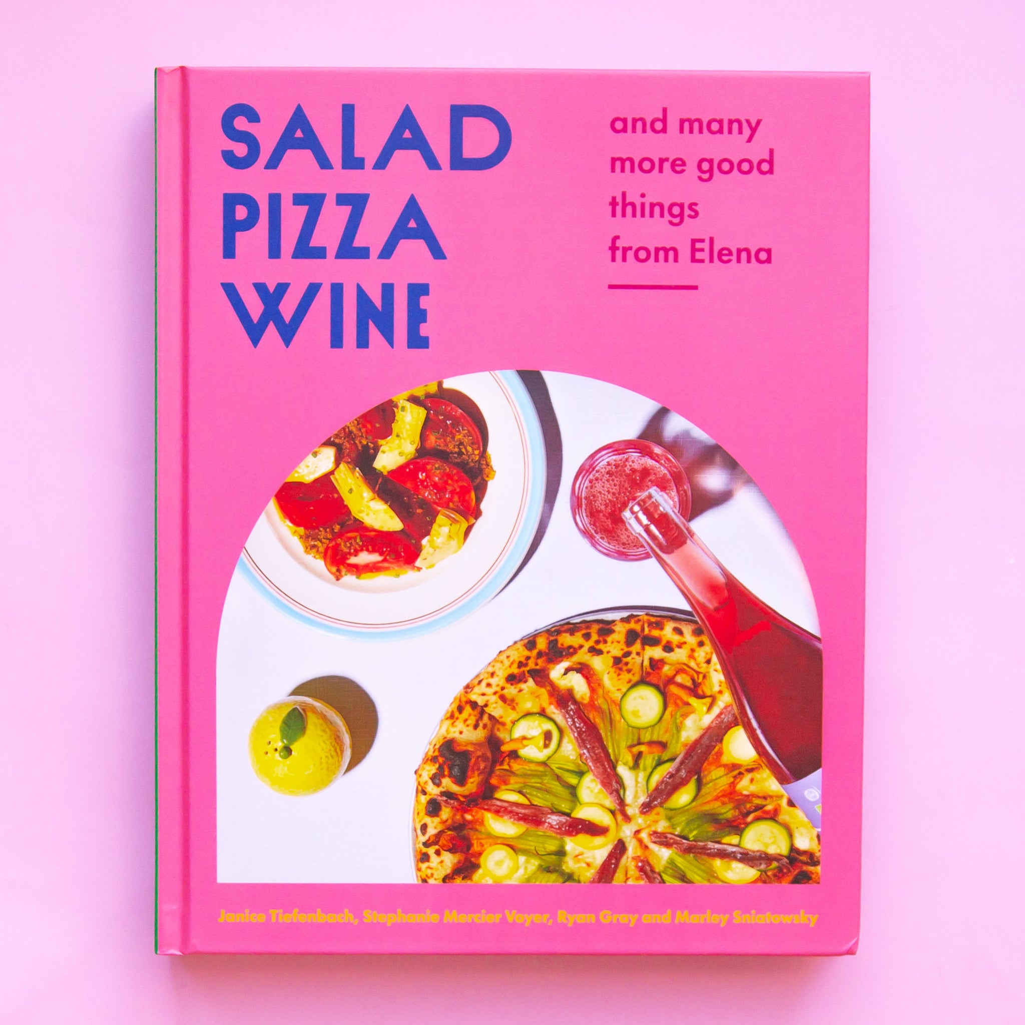On a pink background is a pink hardback book with blue text on the top left corner that reads, &quot;Salad Pizza Wine&quot;, &quot;and many more good things from Elena&quot; along with an arched photo of food on a table scape. 